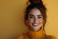 Young Caucasian woman with bright blue eyes and messy bun hairstyle wearing yellow knit turtleneck sweater smiling genuinely against golden background.