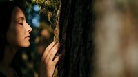 Young woman with dark hair touching tree bark in peaceful forest setting during golden hour sunlight creating serene nature connection moment.