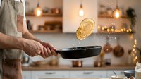 Unrecognizable male chef in linen apron tosses pasta in skillet with flying noodles in modern kitchen with warm lighting and bokeh background.