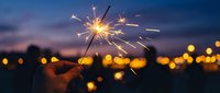 Silhouette of person holding lit sparkler against twilight sky with blurred golden bokeh lights in background, suitable for celebration, festive events, New Year, and holiday marketing materials.
