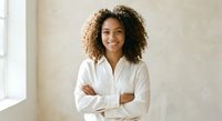 Young African American businesswoman with curly hair smiling confidently with crossed arms in white shirt standing in modern office space.