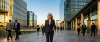 Confident middle-aged woman with blonde hair wearing dark business suit walking through modern urban plaza surrounded by contemporary glass and steel buildings during golden hour, ideal for corporate and professional concepts.