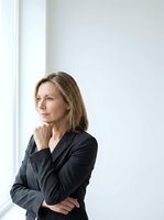 Middle-aged woman with shoulder-length brown hair wearing dark blazer, hand to chin in thoughtful pose near window, suitable for corporate and business concepts.