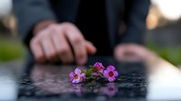 Small pink flowers on dark reflective surface with blurred hands in background creating moody atmospheric composition with shallow depth of field.