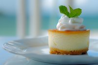 Mini cheesecake topped with whipped cream and fresh mint leaf on white plate. Creamy dessert with graham cracker crust photographed with soft focus background.