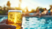 Cold glass of light beer with bubbles held against blurred swimming pool and sunset background, refreshing summer drink by poolside creates vacation mood.