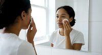 Young African American woman applying skincare product with cotton pad while looking in bathroom mirror during morning routine.