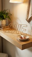 Gold wire LOVE sign on a wooden floating shelf with rose petals in a heart dish, green plant, and warm lamp light creating cozy home decor.