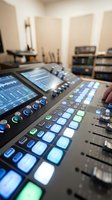 Professional audio mixing console with illuminated buttons and faders in a recording studio control room with acoustic treatment and equipment racks.