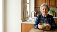 Senior woman with gray hair and light skin tone, wearing a dark patterned sweater, holding a bowl of salad in a modern kitchen with wooden cabinets and white countertops. Healthy eating concept.