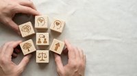 Hands arranging wooden blocks with corporate social responsibility icons including scales, people, trees and gears on neutral background.