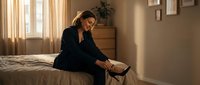 Professional businesswoman in black suit sitting on bed removing high heels after long workday in warm sunlit bedroom interior.