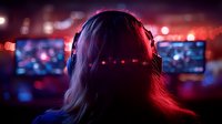 Young female gamer in headphones silhouetted against neon red and blue lighting at esports event, with blurred gaming monitors and crowd in background.