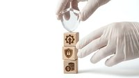Medical professional in white gloves stacking wooden blocks with healthcare symbols including stethoscope, shield and pills on clean background.