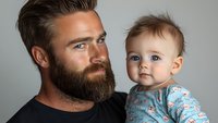 Bearded man in black t-shirt holding adorable baby wearing blue floral pajamas against gray background, emotional family portrait showing tender connection.