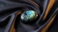 Polished labradorite gemstone with iridescent blue green flash resting on luxurious dark silk fabric for jewelry design and spiritual healing concepts.
