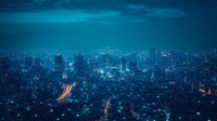 Aerial view of metropolitan cityscape at night with glowing streets and skyscrapers under dramatic blue sky, modern urban landscape with illuminated buildings and highways.