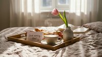 Cozy morning breakfast in bed with coffee cup, croissant and pink tulip flower on wooden tray creating romantic atmosphere for special occasions.
