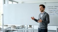 Young adult male with light skin tone and dark hair, wearing business casual attire, presenting to classroom or training session. Educational setting with whiteboard displaying German language content and mathematical formulas.