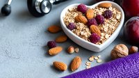 Healthy breakfast with oatmeal, almonds, dried raspberries in heart shaped bowl. Fresh plums, dumbbells on gray background. Fitness and diet concept.