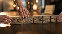 Business strategy concept with wooden blocks spelling start plan work check win arranged by hands on office desk with warm lighting atmosphere.