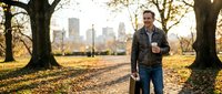 Man jogging through urban park in autumn with city skyline visible in background. Male runner in casual athletic wear exercising outdoors during fall season for fitness and wellness.