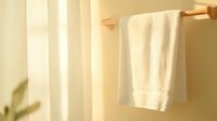 Soft white towel hanging on wooden rack against beige wall with gentle morning sunlight casting shadows, creating minimalist bathroom atmosphere.