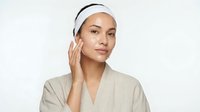 Young woman applying skincare cream to face wearing white headband and beige robe for daily beauty routine and facial care regimen.