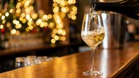 White wine being poured into a glass on a wooden bar counter with warm golden bokeh lights and festive holiday decorations in the background, suitable for beverage, hospitality and celebration content.