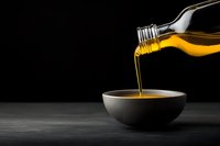 Golden olive oil pouring from glass bottle into dark ceramic bowl on black background, dramatic lighting creates luxurious culinary atmosphere.