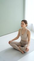 Young Caucasian woman in beige activewear practicing meditation in lotus pose on yoga mat in bright minimalist studio with natural light.