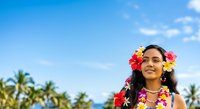 Woman with dark hair and South Asian ethnicity wearing vibrant floral lei and flower crown against tropical palm tree backdrop and blue sky. Perfect for travel, vacation, and cultural celebration content.