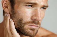 Caucasian male model with stubble beard touching neck in contemplative pose against light background, showcasing masculine grooming and skincare concept.