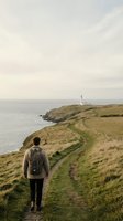 Hiker with backpack walking on coastal trail toward lighthouse on grassy headland overlooking calm sea, adventure and outdoor exploration.