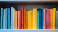 Colorful books arranged on wooden shelf in rainbow order, spines showing vibrant hues of blue, orange, yellow, pink and red creating modern minimalist library display.