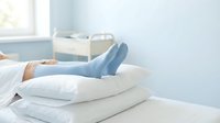 Person with light skin tone wearing blue socks resting legs on stacked white pillows in bright bedroom with bed and window, conveying relaxation and comfort for wellness and lifestyle content.