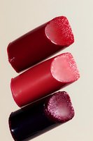 Diagonal arrangement of three lipstick bullets in red and burgundy shades with frost effect texture on cream background, showcasing cosmetic color variety.