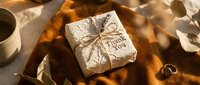 Rustic thank you gift box wrapped in textured paper with twine bow, placed on warm golden fabric with dried leaves and rings.