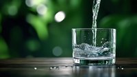Fresh clean water pouring into transparent glass on wooden surface with natural green bokeh background for healthy lifestyle and hydration concepts.