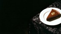 Slice of rich chocolate cake with gold leaf garnish on white plate against dark textured surface, suitable for bakery, dessert, and food photography projects.
