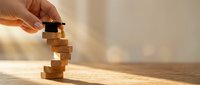Hand carefully placing wooden block on unstable tower demonstrating balance, risk management and strategic decision making in business planning.