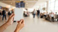 Hands holding blank white tablet device in busy airport terminal with travelers and departure board visible in background.