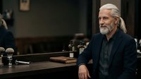 Mature man with long gray hair and beard, Caucasian ethnicity, wearing dark button-up shirt, seated at conference table during professional meeting or interview setting.