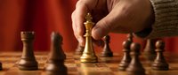 Hand moving golden chess king piece on wooden board during strategic game with warm lighting and red background creating focused atmosphere.