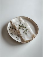 Elegant table setting with white linen napkin decorated with delicate green sprigs and scattered black seeds on ceramic plate against light background.