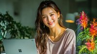 Young Asian woman smiling while working on laptop in modern home office with colorful plants creating productive workspace atmosphere for remote work and freelancing.