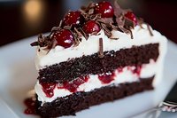 Decadent chocolate cake slice layered with fresh cream and cherries, topped with chocolate shavings and glazed cherries on white plate, close-up view with soft focus.