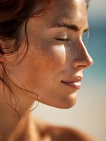Close-up profile portrait of young woman with freckles and natural glowing skin in warm sunlight against blurred beach background, eyes closed in peaceful expression.