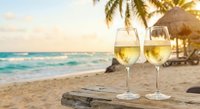 Two glasses of white wine on wooden table at tropical beach resort with palm trees and ocean waves during golden hour sunset for romantic vacation celebration.
