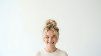 Blonde woman with hair styled in top bun, wearing cream ribbed sweater, smiling at camera against white background. Portrait for business, lifestyle, and wellness content.